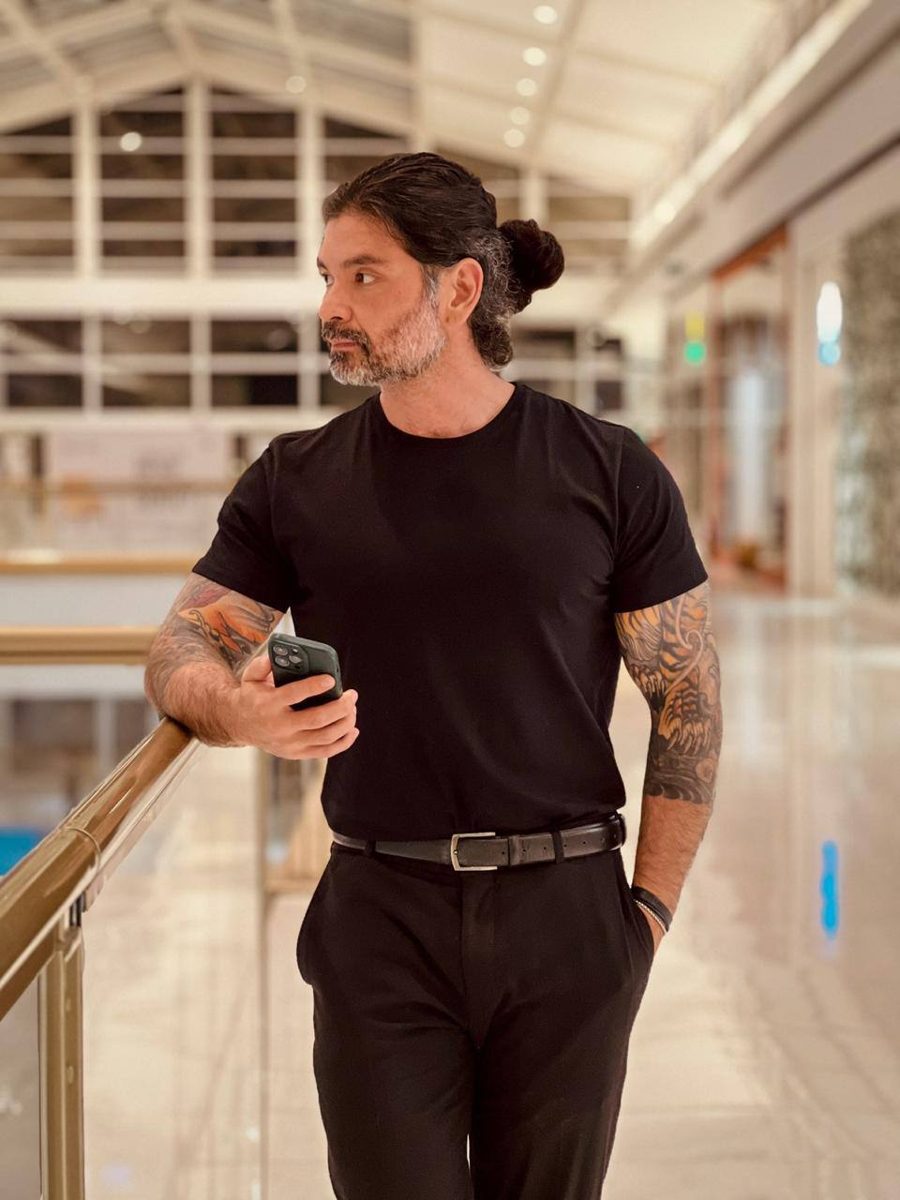 Bruno Nakabashi de camiseta e calça pretas, apoiado no corrimão de um corredor claro, olhando para o lado